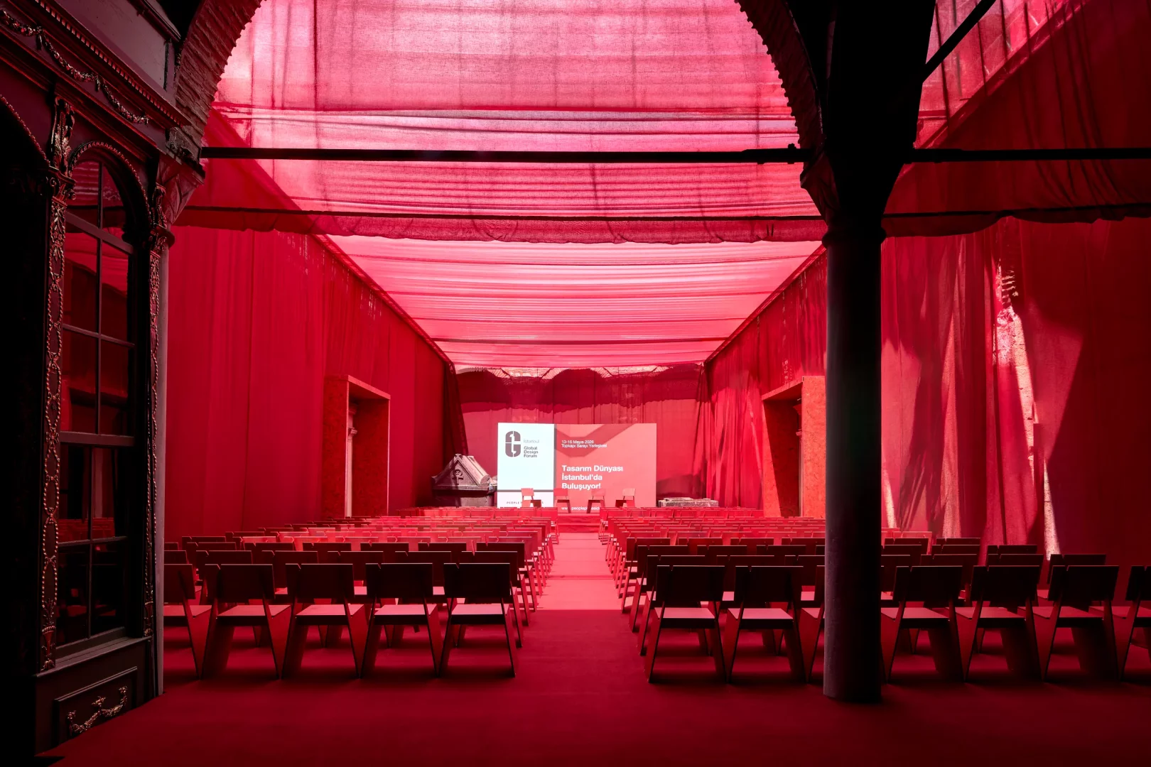 13-16 Mayıs 2026 Global Design Forum İstanbul etkinliğinin görseli, Tasarım Dünyası İstanbul'da Buluşuyor! yazılı ekranın önünde, tavanı ve duvarları kırmızı kumaşlarla kaplı, sahneye dönük düzenlenmiş boş kırmızı sandalyelerle dolu, tasarımı şehrin farklı noktalarına yayma vizyonunu yansıtan özel bir etkinlik kurgusunu gösteriyor.