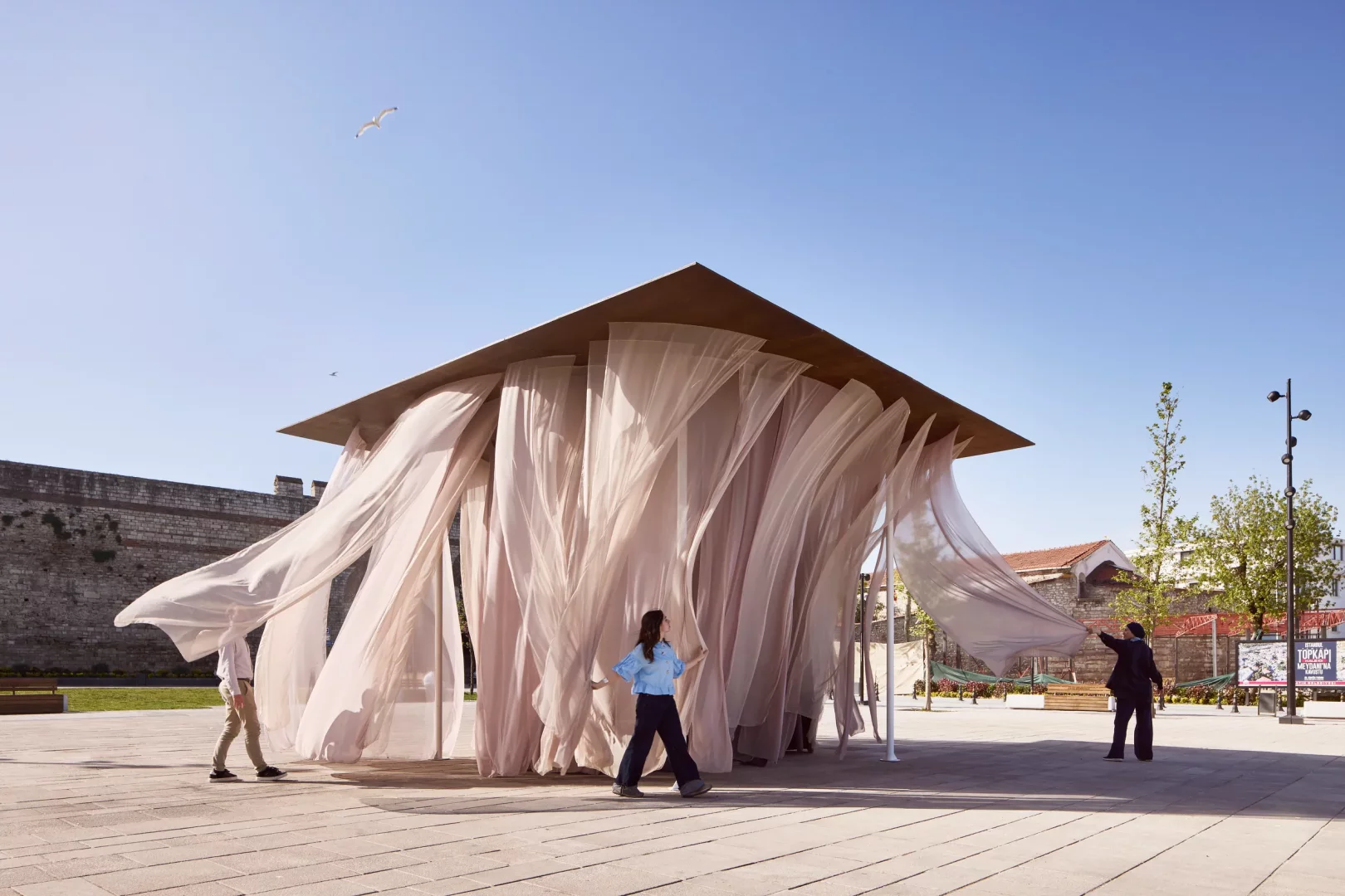 Global Design Forum İstanbul kapsamında, Waugh Thistleton Architects imzalı, rüzgarda uçuşan şeffaf bej kumaş perdelerle çevrili, kare çatılı Pavilion of the Moment adlı kamusal yerleştirmede insanlar dolaşırken, eserin geçicilik temasını vurgulayan mimarisi tarihi bir şehir duvarının önünde parlıyor.