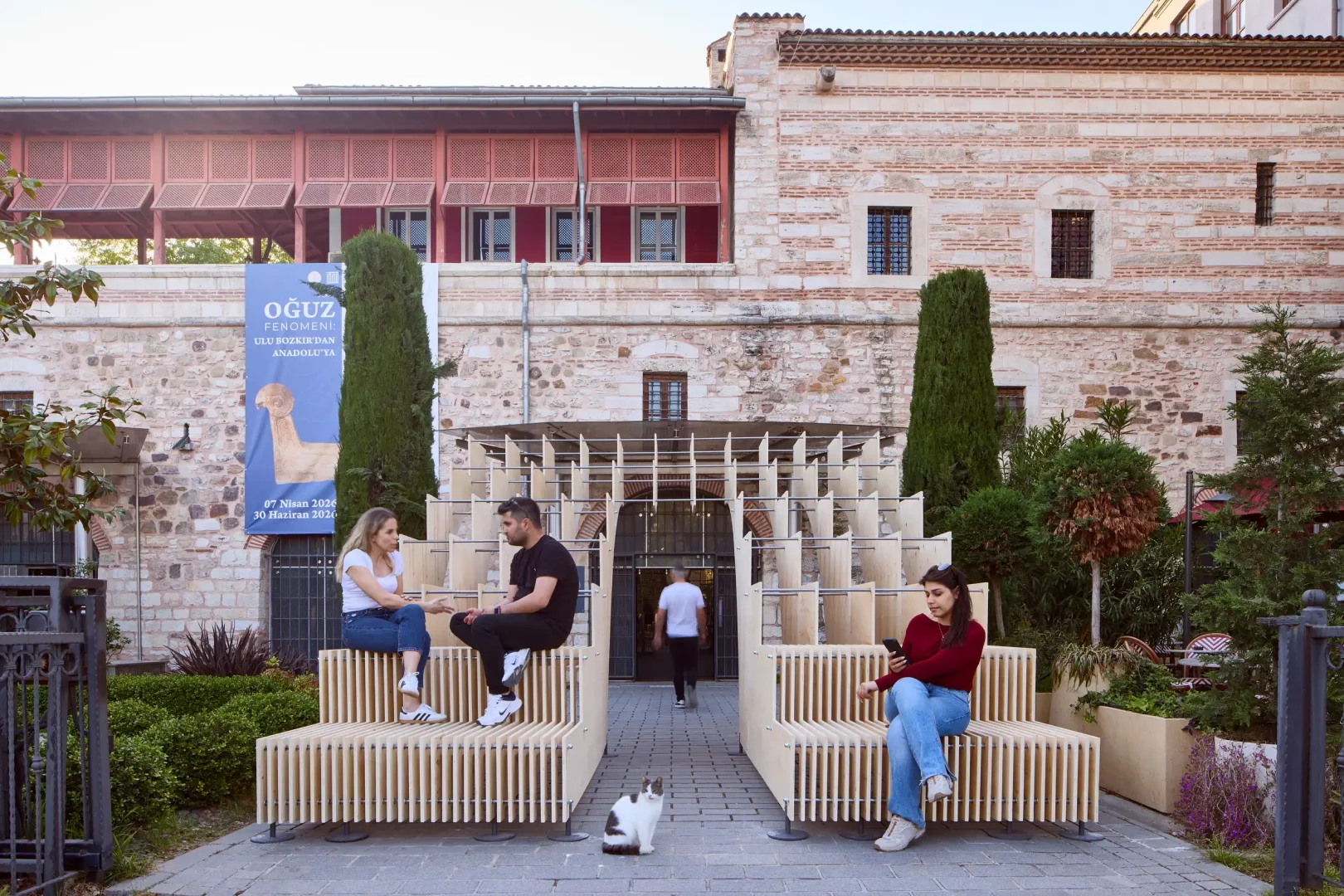 Tarihi taş bir yapının bahçesinde konumlanan, Global Design Forum İstanbul'un tasarımı şehre yayma fikrini yansıtan, Waugh Thistleton Architects imzalı ahşap ve geçici bir kamusal yerleştirme. Modüler banklarında sohbet eden ve telefonuna bakan insanlarla birlikte, ön planda oturan bir kedi, modern tasarımın tarihi doku ve gündelik yaşamla buluştuğu samimi bir anı betimliyor.