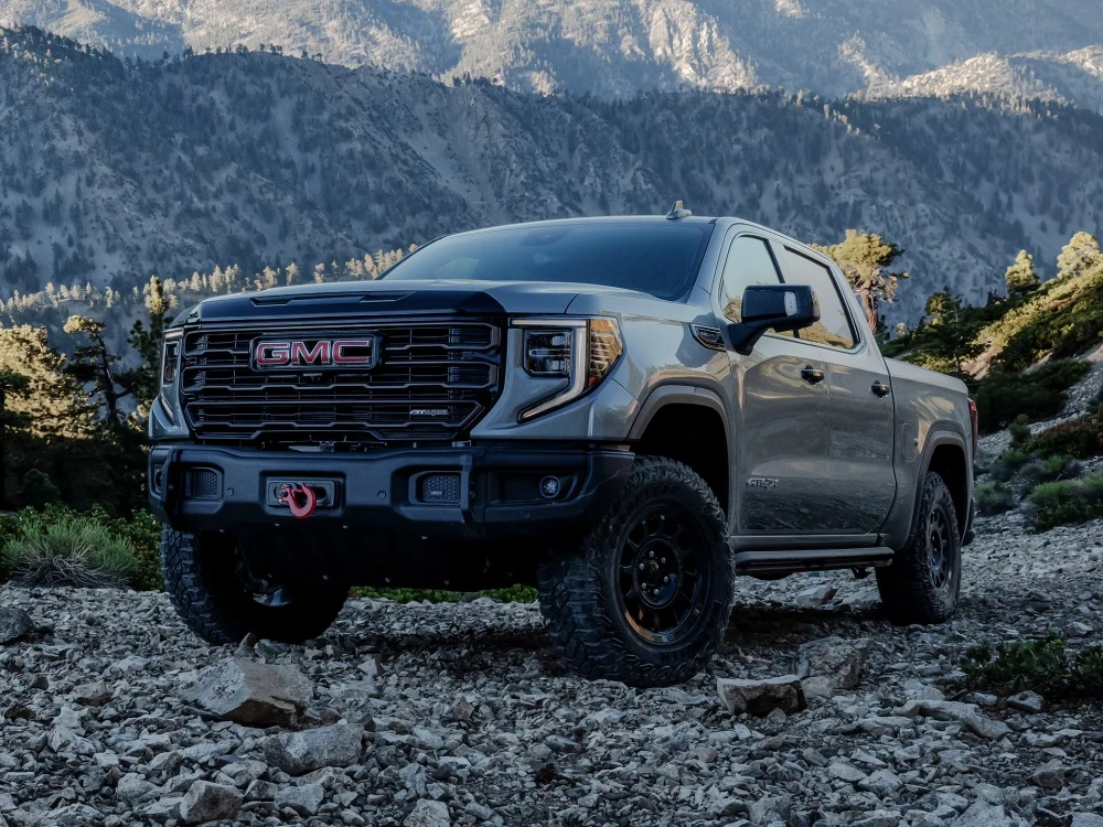 Engebeli, kayalık bir arazi üzerinde konumlanmış, ön kısmında kırmızı bir çekme kancası bulunan gri renkli GMC Sierra AT4X pickup, dağlık arka planıyla off-road macerasına hazır bir görünüm sunuyor.