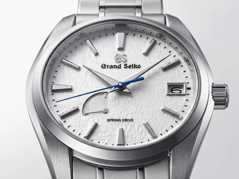 Grand Seiko Snowflake kol saati, kendine özgü kar tanesi dokulu beyaz kadranı, mavi saniye ibresi, 3 yönündeki tarih penceresi ve 8 yönündeki güç rezervi göstergesiyle Japon saatçiliği estetiğini ve Spring Drive teknolojisini yansıtır.