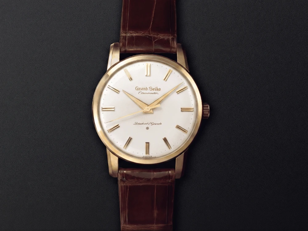 Siyah mat bir zeminde, kahverengi timsah derisi desenli kayışa sahip, altın renkli kasası ve altın çubuk indeksli beyaz kadranı olan, Grand Seiko Chronometer yazısı belirgin bir vintage Japon saati yakından gösteriliyor.