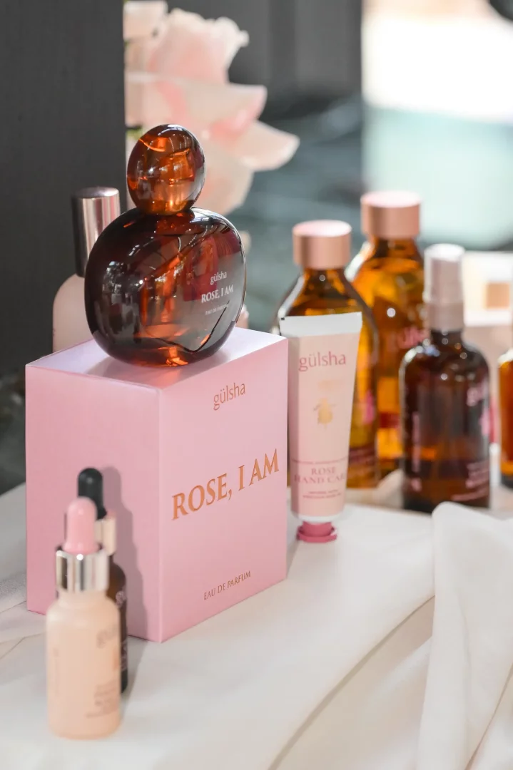 gülsha ROSE, I AM lansmanı için özel olarak tasarlanmış, koyu kehribar rengi, yuvarlak kapaklı Eau de Parfum şişesi pembe kutusu üzerinde dikkat çekerken, arka planda markanın gül temalı el kremi ve çeşitli damlalıklı serum şişeleri gibi ürün yelpazesinin diğer parçaları zarifçe sergileniyor.