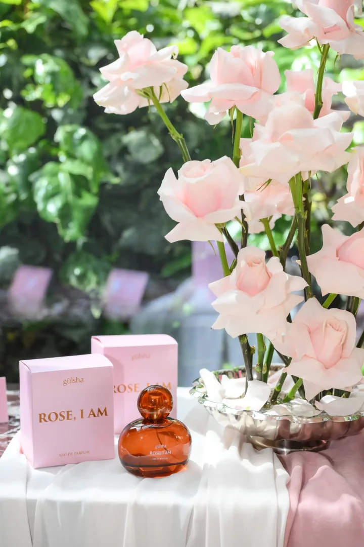 gülsha Rose, I Am lansmanından bir karede, kehribar rengi yuvarlak şişesi ve iki adet açık pembe kutusu beyaz örtülü zarif bir masada, arka planda yemyeşil bitki örtüsü ve gümüş bir kase dolusu canlı açık pembe güllerle birlikte şık bir şekilde sergileniyor.
