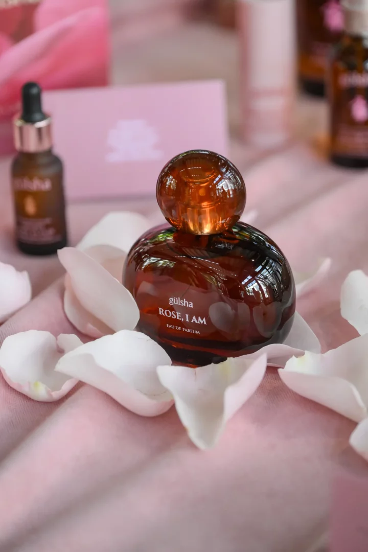 Pembe bir zemin üzerinde, beyaz gül yapraklarıyla süslenmiş ortamda, ön planda yuvarlak hatlara sahip koyu kehribar renkli gülsha ROSE, I AM EAU DE PARFUM şişesi göz alıcı bir şekilde konumlanmış; arka planda ise markanın lansman etkinliğini çağrıştıran diğer ürünler ve ambalajlar flu bir şekilde seçilmektedir.
