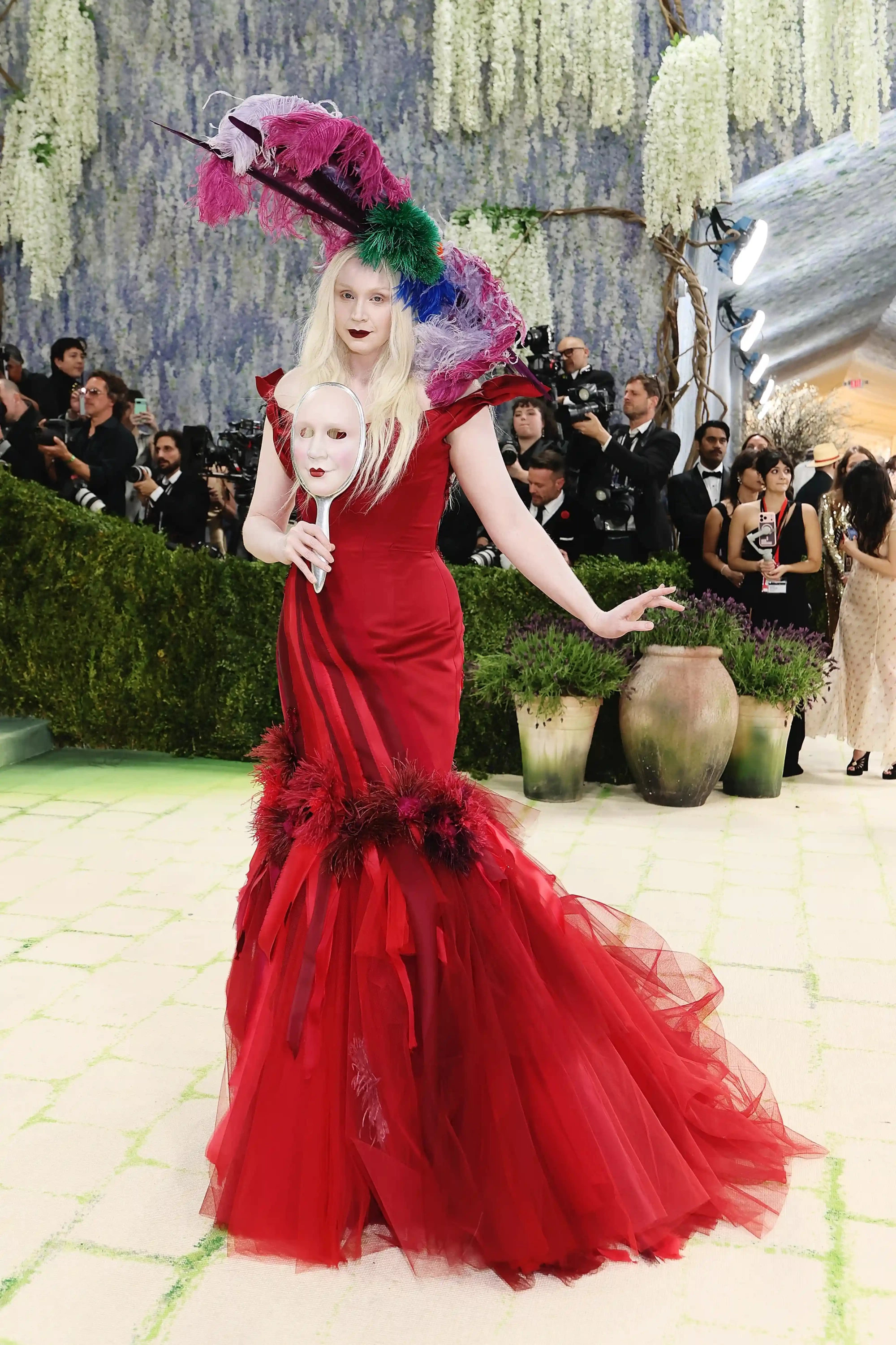 Gwendoline Christie, 2026 Met Gala kırmızı halısında, heykelsi duruşuyla parlak kırmızı, tüylü bir elbise, gösterişli çok renkli bir başlık ve yüz maskesiyle Fashion Is Art temasına uygun, iddialı bir kostüm sanatı sergiliyor.