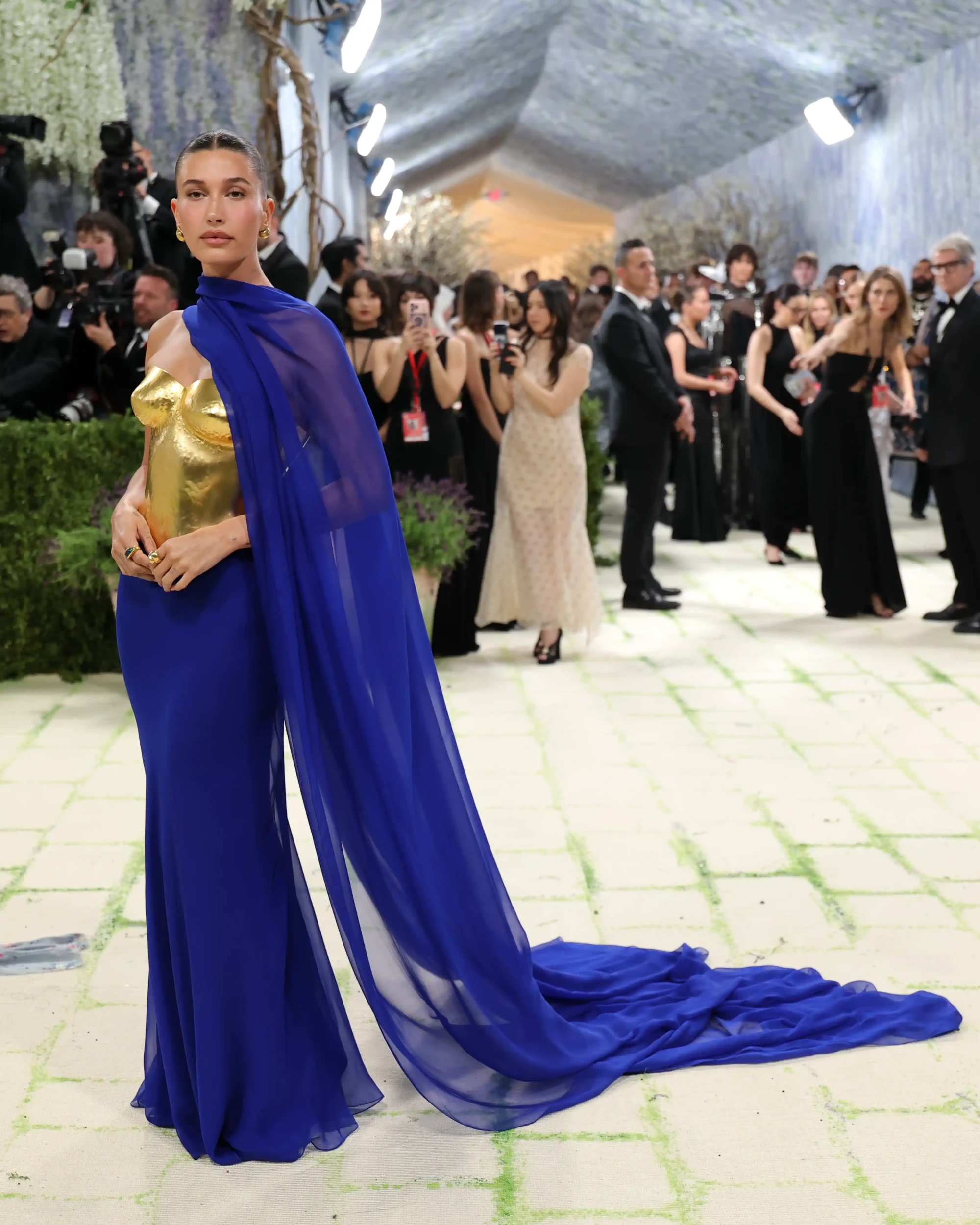 Hailey Bieber, 2026 Met Gala kırmızı halısında, altın rengi heykelsi büstiyeri ve arkasına doğru akıcı bir şekilde uzanan uzun kraliyet mavisi peleriniyle, moda ve sanatın uyumunu sergileyen büyüleyici bir kostüm içinde objektiflere poz veriyor.