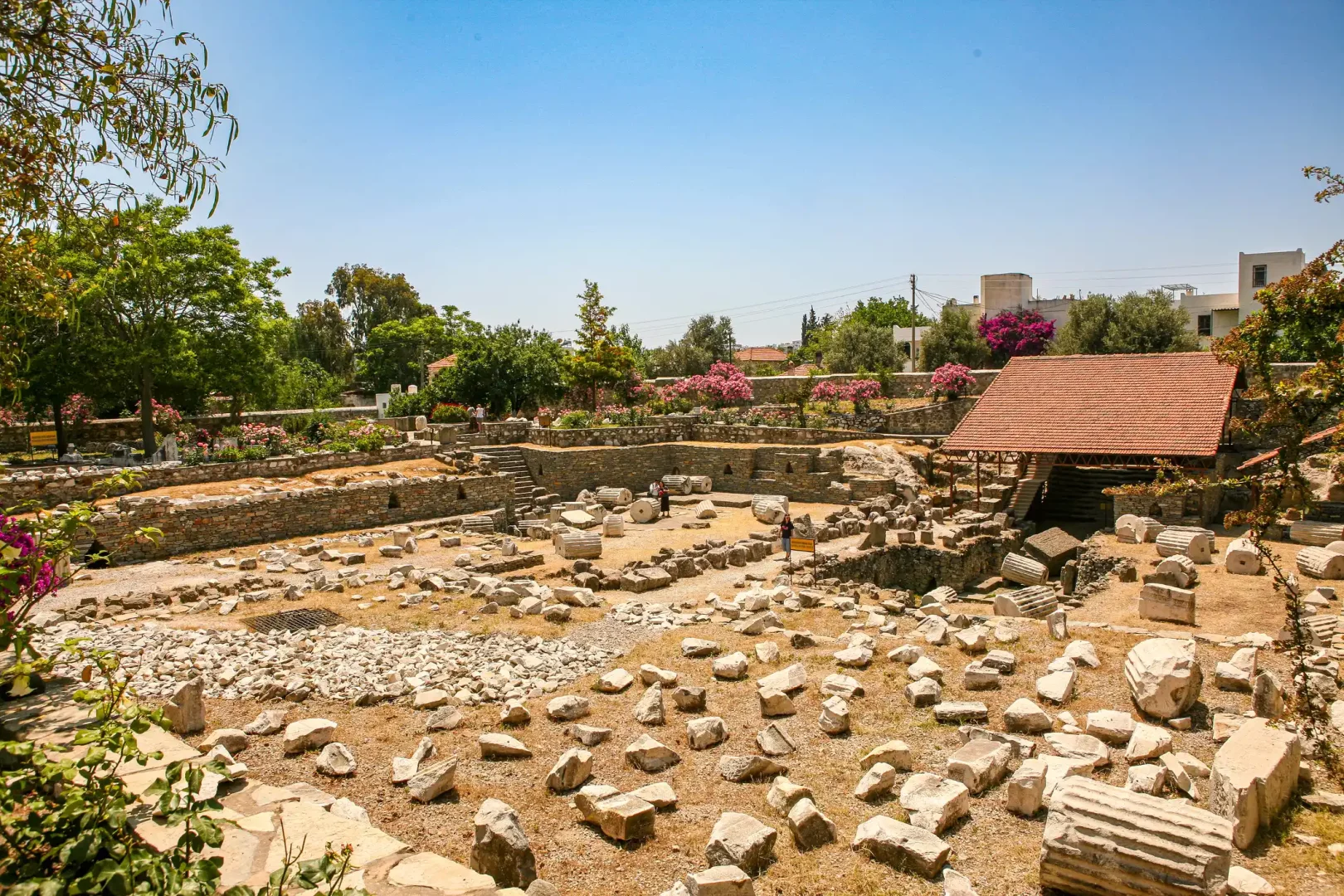 Halikarnas Mausoleum alanı kalıntıları, güneşli bir günde, etrafa dağılmış mermer sütun parçaları, taş bloklar ve kısmi duvar yapıları ile antik bir geçmişi sergiliyor; sağda ahşap çatılı bir kazı koruma alanı, arka planda yeşil ağaçlar, renkli çiçekler ve modern yapılarla çevrili.