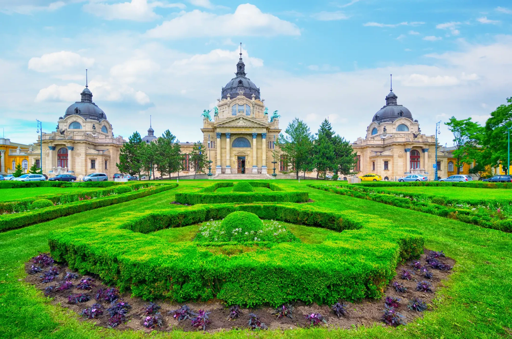 Budapeşte'deki Széchenyi Termal Hamamı, yemyeşil çitler ve mor çiçeklerle bezeli geniş bahçesi önünde yükselen, görkemli kubbeleri ve süslü bej cephesiyle parlak mavi gökyüzü altında tüm ihtişamını sergiliyor.