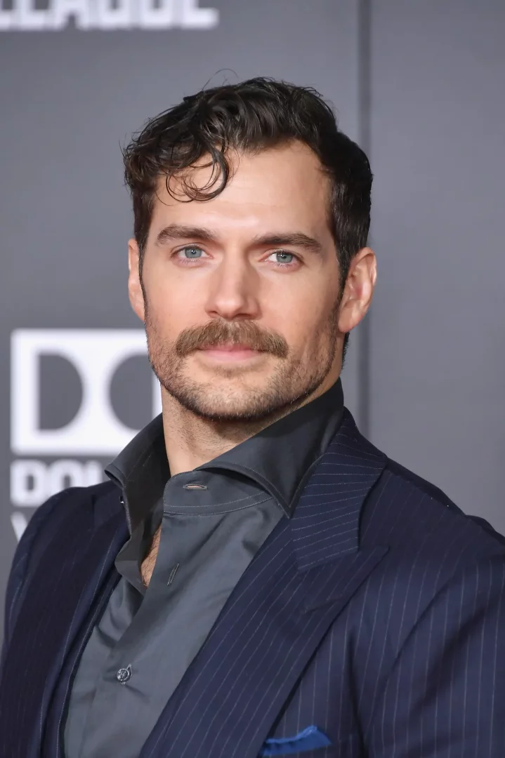 Ünlü oyuncu Henry Cavill, karakteristik kıvırcık koyu saçları, dikkat çekici mavi gözleri, şık bıyığı ve kısa sakallarıyla, lacivert çizgili takım elbisesi ile kamera karşısında dururken çekilmiş bu yakın planda karizmatik bir duruş sergiliyor.