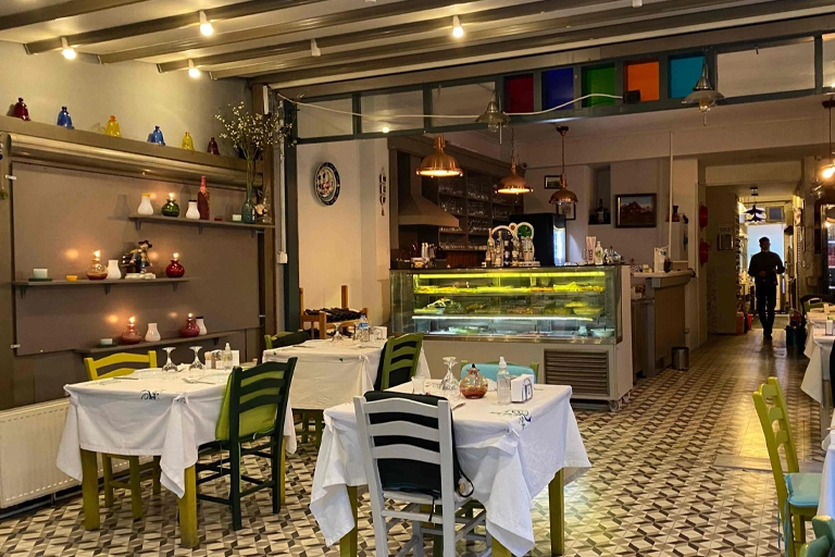 Heybeliada'da yer alan Heyamola Ada Lokantası'nın içinden çekilmiş bu geniş açılı fotoğraf, duvar raflarındaki renkli cam objeler ve yanan mumlarla aydınlatılmış sade masa düzenleri, bar tezgahındaki mezeler ve baklava desenli yer karolarıyla sıcak ve davetkar bir atmosfer sunuyor.