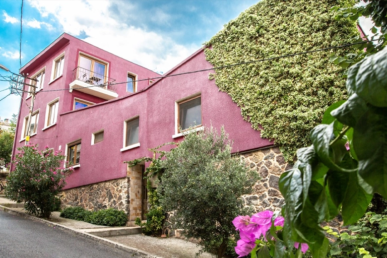 Heybeliada'daki Lisola Guesthouse otelin begonviller ve yeşilliklerle çevrili, kısmen sarmaşıklarla kaplı, taştan bir tabana sahip, canlı mor renkli ve üst katında balkonu bulunan dış cephesi, bulutlu mavi gökyüzü altında yokuşlu bir sokaktan görülüyor.