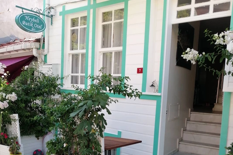 Heybeli Butik Otel tabelasının asılı olduğu, beyaz cephesini turkuaz çerçeveli pencereler ve yeşil bitkilerin çevrelediği şirin bir giriş, Heybeliada'nın sıcak atmosferini yansıtan bir butik otel deneyimi vadediyor.