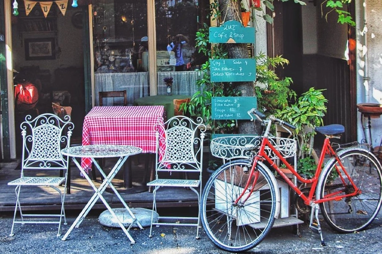 Heybeliada'da bulunan Luz Cafe'nin dış mekanında, beyaz ferforje masa ve sandalyelerin yer aldığı, kırmızı beyaz kareli masa örtüsüyle dikkat çeken bir masanın önünde, menü panolarının asılı olduğu ağacın yanında park etmiş kırmızı bir bisikletle şirin ve davetkar bir atmosfer görülüyor.