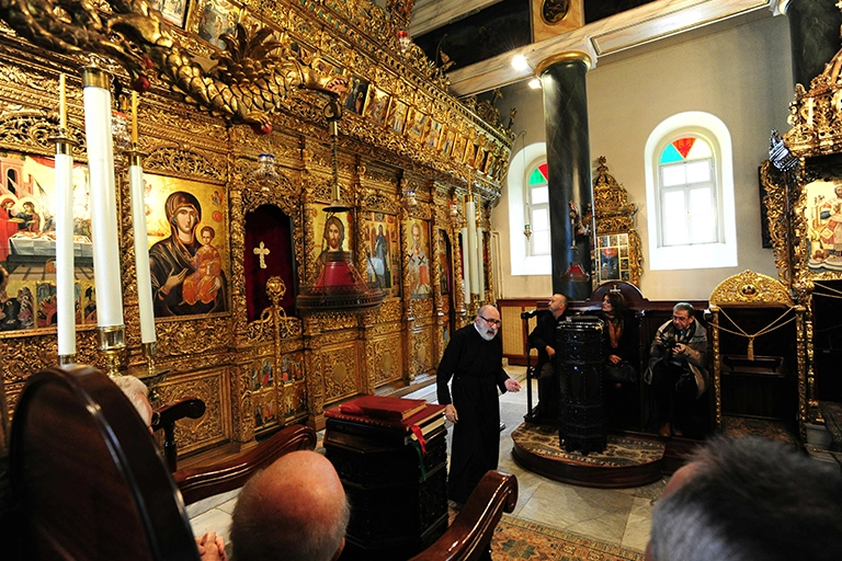 Heybeliada Ruhban Okulu'nun içindeki bir şapelde, altın varaklı ve ikonalarla süslü görkemli ikonostasisin önünde cübbesiyle ayakta duran rahip, etrafındaki cemaate hitap ederken, iki kemerli pencereden ışık süzülen bu tarihi ve ruhani mekanda dinleyiciler tarafından dikkatle izleniyor.