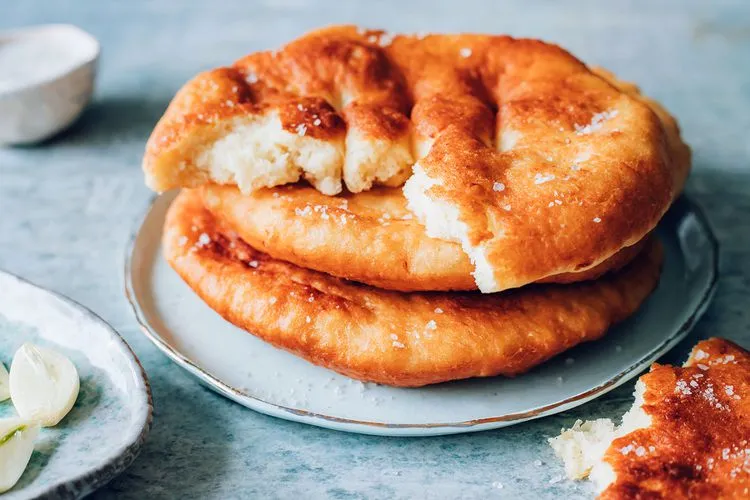Aydınlık bir zeminde, altın renginde kızarmış, puf puf görünümlü, üzerine iri taneli tuz serpilmiş üç adet Macar kızarmış ekmeği (Lángos) üst üste duruyor; en üstteki ekmek ortadan kırılmış, arkada küçük bir kase tuz ve solda soyulmuş sarımsak dişleri görünen iştah açıcı bir sunum.