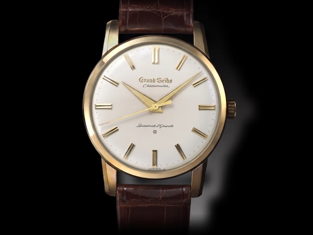 Siyah bir arka plan üzerinde, zarif altın renkli kasası, klasik krem rengi kadranında altın çubuk indeksleri ve akrep yelkovanları ile Grand Seiko Chronometer, Diashock 25 jewels yazıları bulunan, kahverengi timsah desenli deri kayışına sahip, ilk Grand Seiko Japon vintage saat modelinin detaylı ve estetik bir yakın çekimi.