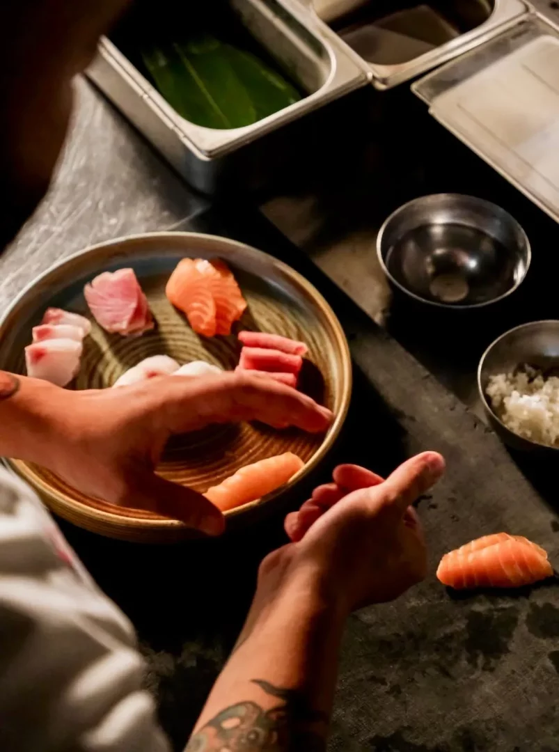 Bir şefin elleri, çeşitli somon, orkinos ve beyaz balık dilimleriyle dolu bir tabağın yanında, pirinci ustaca şekillendirerek özenle bir nigiri hazırlıyor; bu an, kişiselleştirilmiş bir omakase deneyiminin başlangıcını gözler önüne seriyor.
