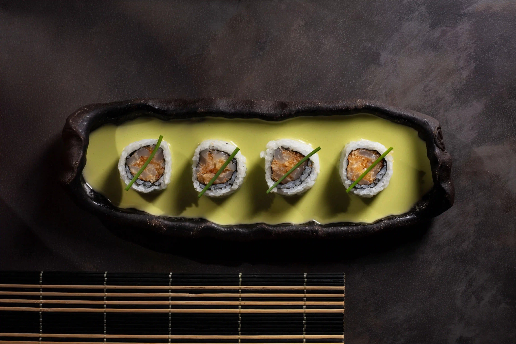 Dört adet beyaz pirinçli, ortası balık dolgulu ve taze frenk soğanı ile süslenmiş sushi rulosu, koyu renkli bir tabakta parlak sarı-yeşil bir sosun içinde özenle sunularak İstanbul’un popüler sushi restoranlarındaki şef imzalı inovatif lezzetleri yansıtıyor.