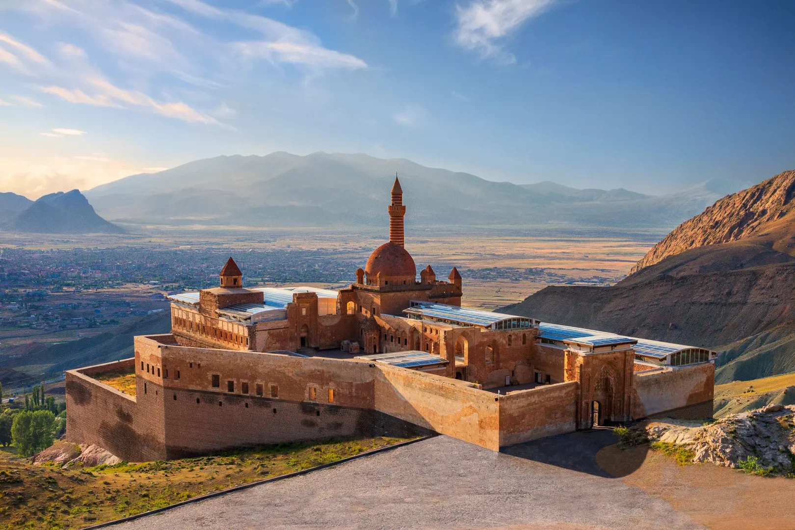 İshak Paşa Sarayı'nın gün batımı ışıkları altında parlayan, kırmızımsı taştan yapılmış kubbeli camisi, sivri minaresi ve geniş avlularıyla bir tepenin yamacına kurulmuş görkemli kompleksi, aşağıda uzanan şehri ve uzaklardaki dağları gösteren nefes kesici bir panoramik manzarayı gözler önüne seriyor.