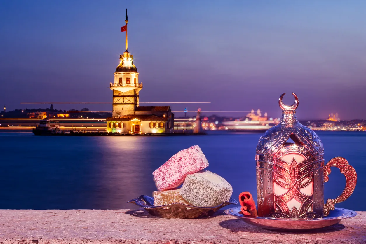 Gün batımında mor ve mavi tonlarıyla aydınlanan Kız Kulesi'nin eşsiz İstanbul manzarası önünde, geleneksel işlemeli bir fincan ve çeşitli renklerde sunulan lokumlar, şehrin ikonik tatlarını ve kültürel atmosferini yansıtan bir Tadım Sineması deneyimine gönderme yapıyor.
