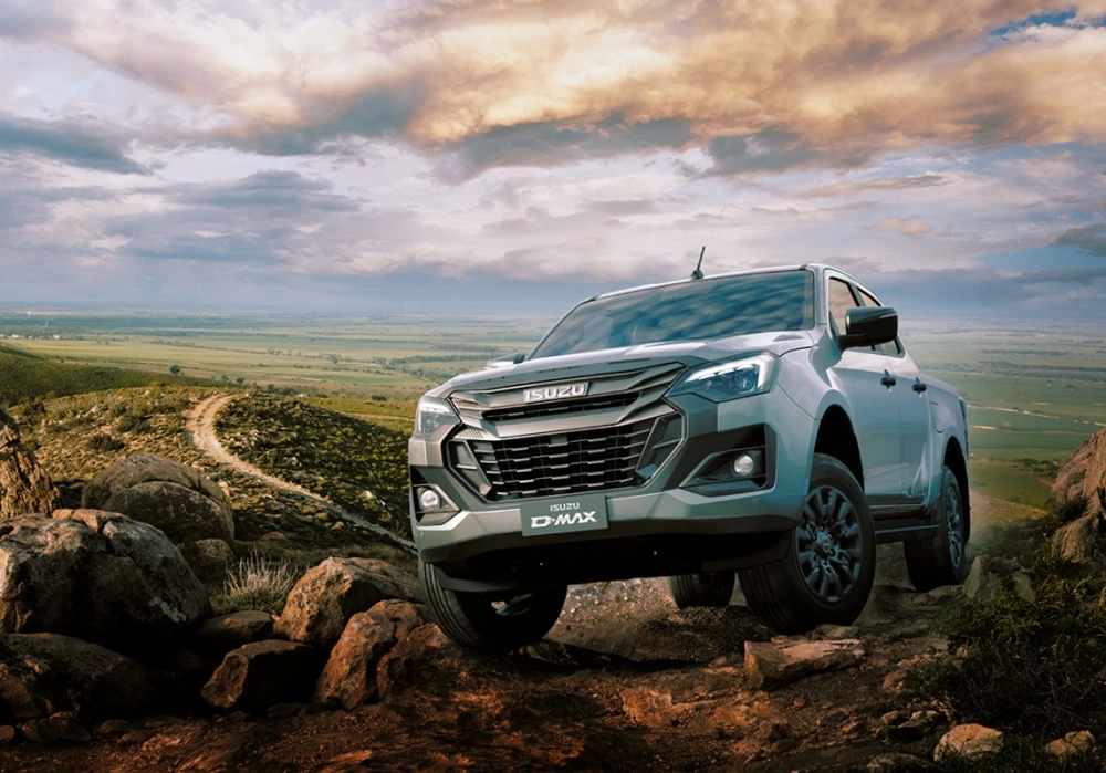 Ön planda gri renkli, dayanıklı bir Isuzu D-MAX pickup kamyonet, zorlu kayalık ve engebeli bir arazide offroad sürüşü yaparken, arka planda geniş yeşil ovalar ve dramatik bulutlarla dolu gökyüzü manzarasıyla güçlü bir duruş sergiliyor.