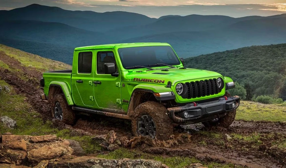 Parlak yeşil renkli Jeep Gladiator Rubicon pickup, çamurlu ve engebeli bir offroad patikasında ilerlerken, yerden yüksek yapısı ve arazi lastikleriyle zorlu koşullara meydan okuyor, arkada ise dağlık ve ormanlık bir manzara uzanıyor.