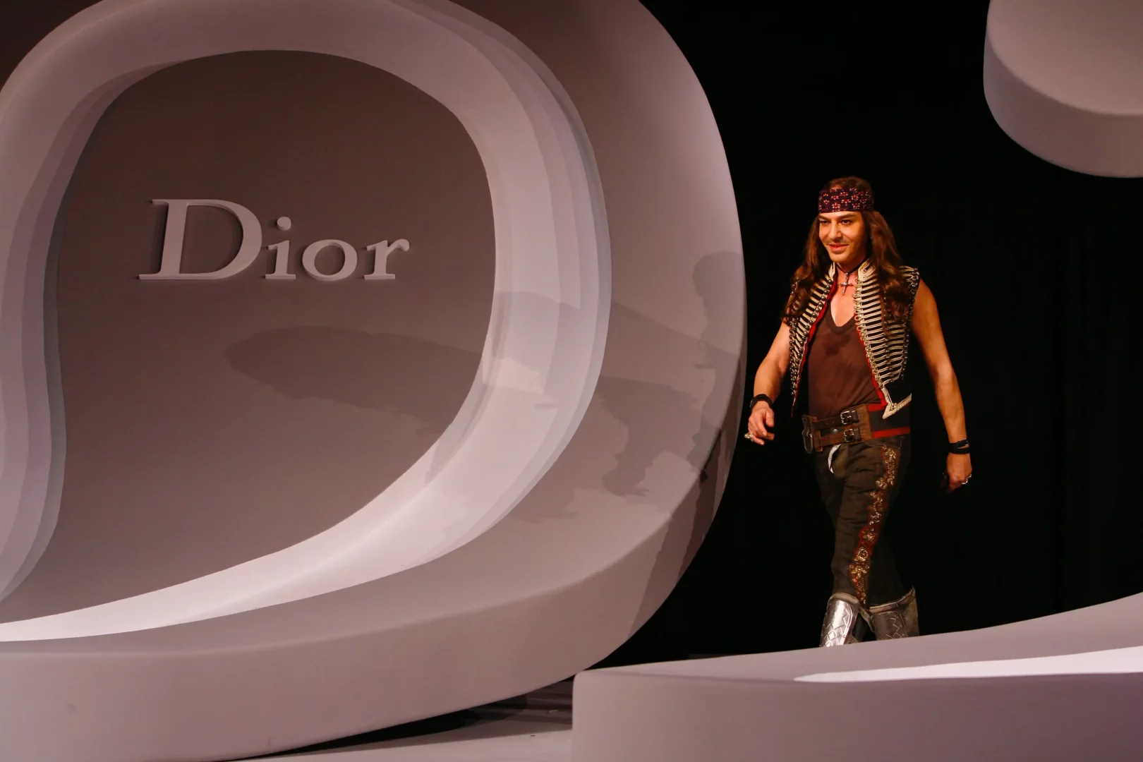 Uzun saçları, desenli bandanası, askeri esintili yeleği ve işlemeli pantolonuyla podyumda yürüyen John Galliano, arkasında büyük bir Dior logosuyla moda ve tasarım dünyasının Met Gala gibi etkinliklerdeki yaratıcı ruhunu yansıtıyor.