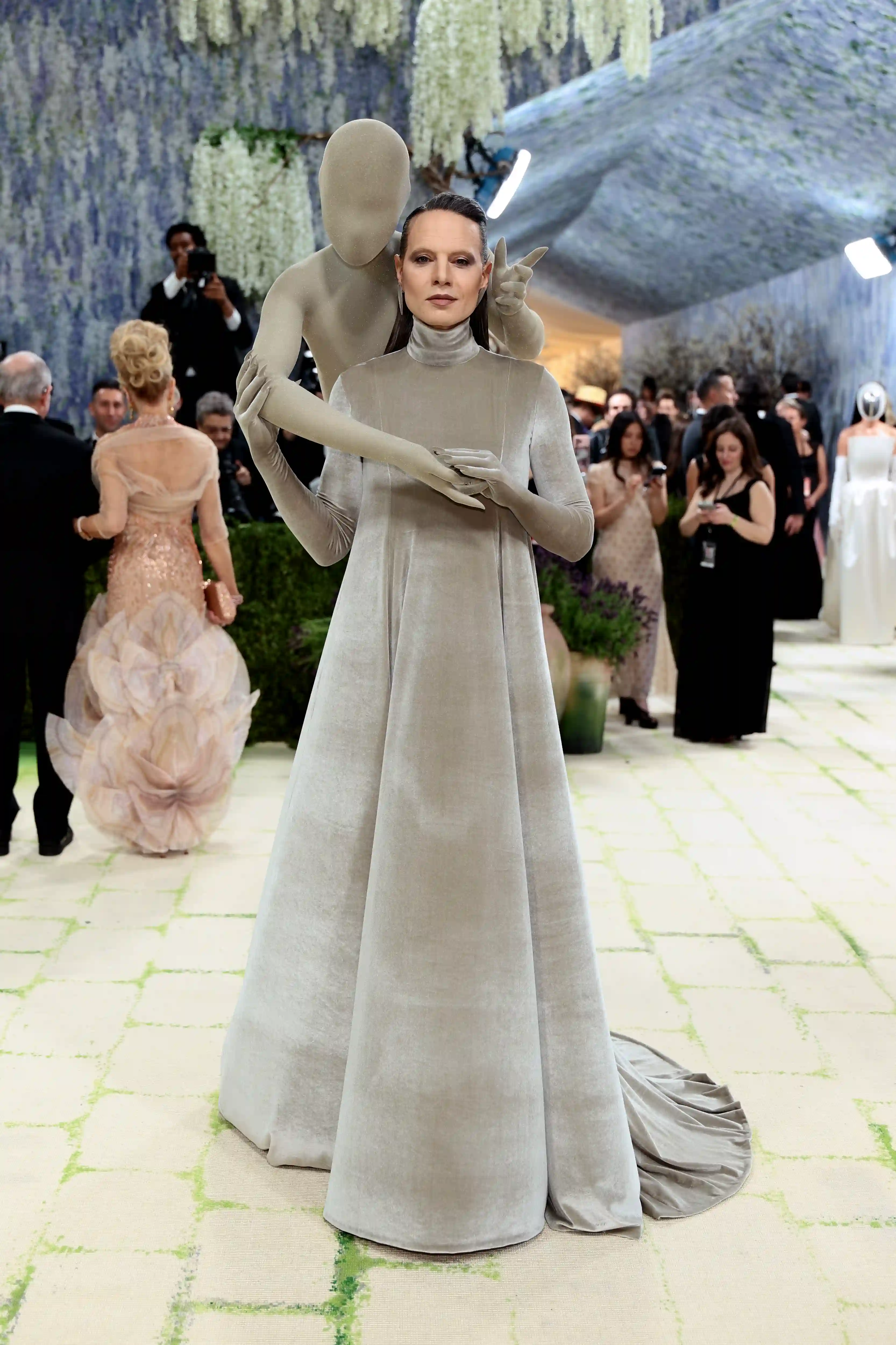 Uzun, boğazlı, açık gri kadife elbisesi içinde, arkasındaki heykelsi manken figürüyle bütünleşen bir kadın, Met Gala'nın kırmızı halısında Fashion is Art temasını modern bir kostüm sanatı yorumuyla sergiliyor.