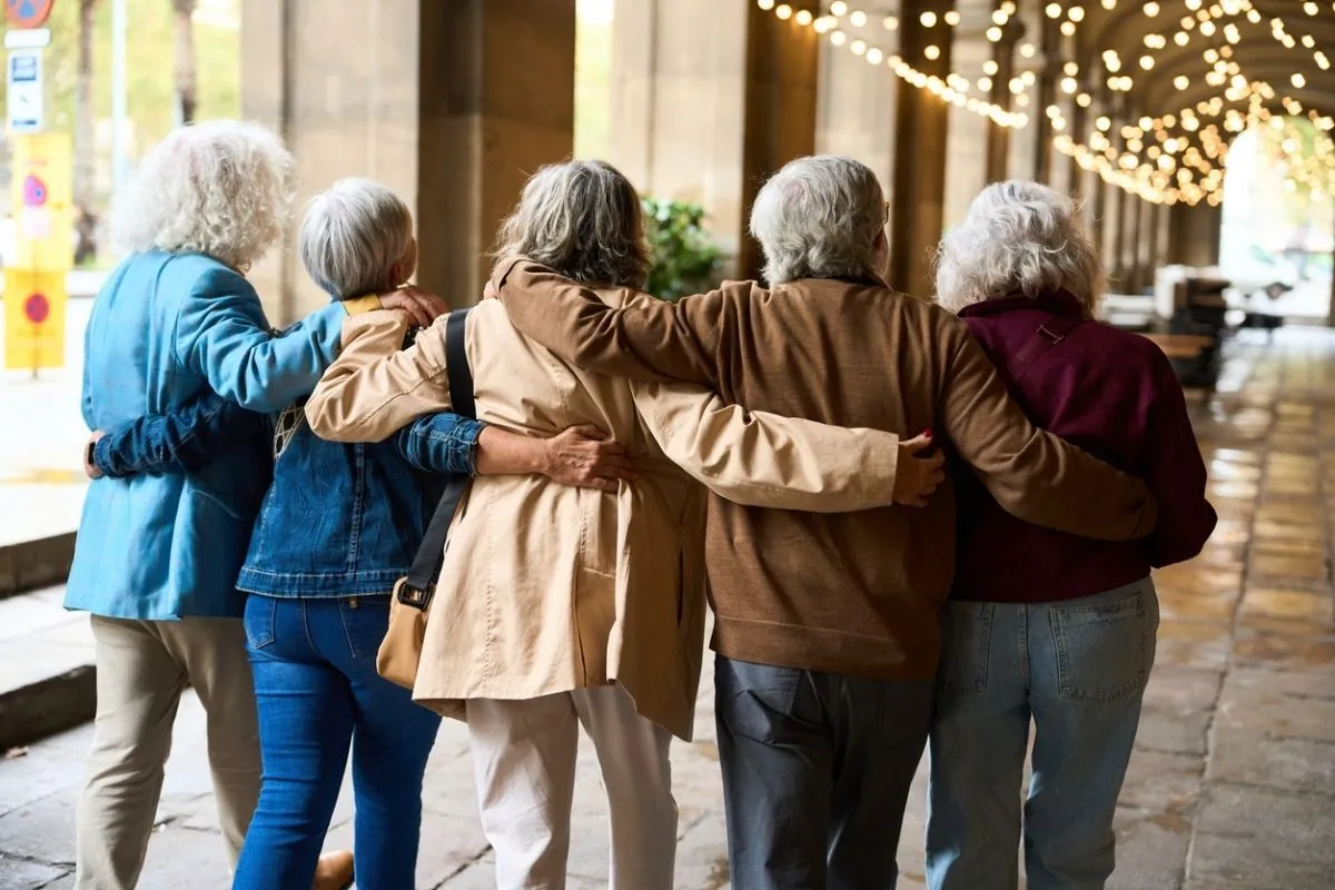 Omuzlarında kolları birbirini saran, arkadan görünen, beyaz saçlı beş yaşlı kadın, ışıklı bir geçidin altında birlikte yürüyerek kadınlar için longevity ve yaşam kalitesi hedefini destekleyen aktif ve güçlü bir sosyal bağı sergiliyor.