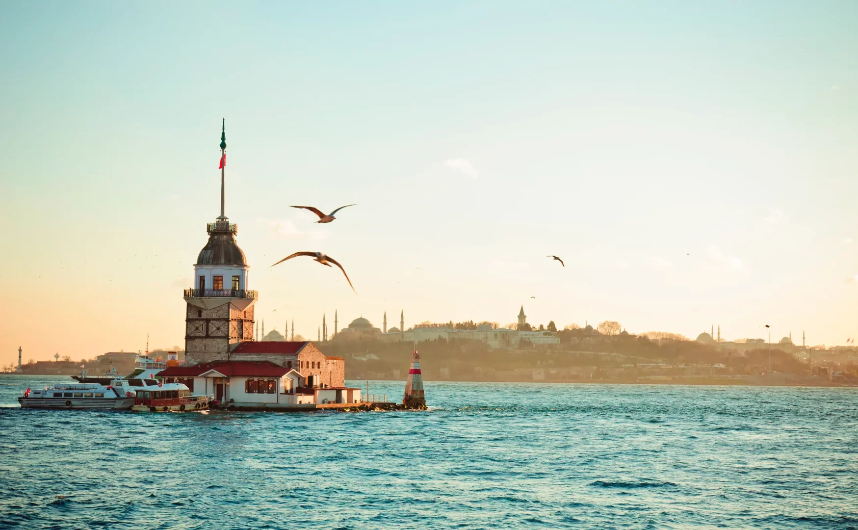 İstanbul Boğazı'nın masmavi sularında yükselen tarihi Kız Kulesi, gün batımının altın rengi ışıkları altında martıların gökyüzünde süzüldüğü, karşı kıyıda İstanbul siluetindeki saray ve cami kubbelerinin belirdiği büyüleyici bir manzara sunuyor.