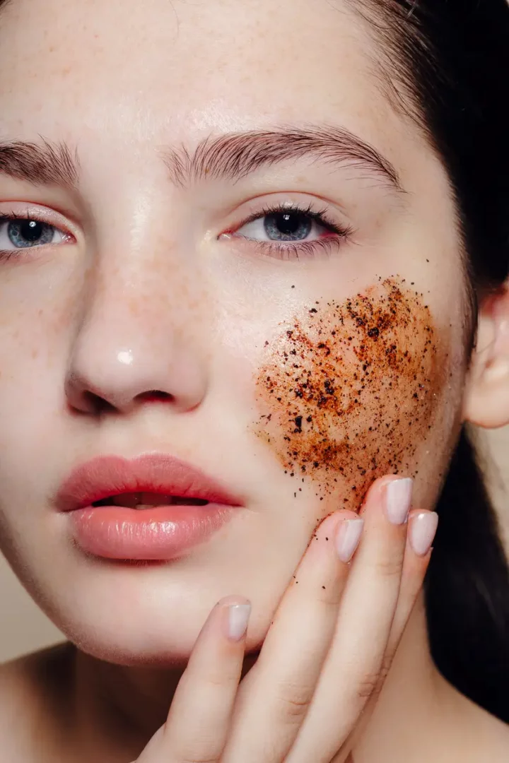 Yakından çekilmiş bu fotoğrafta, çilli ve açık tenli genç bir kadının sol yanağında, kahverengi granüllere sahip bir peeling ürünü bulunuyor ve eliyle cildine nazikçe uygulayarak peeling yaptığını gösteriyor.