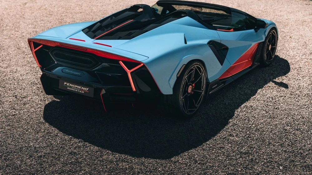 Gökyüzü mavisi ve kırmızı renklerin hakim olduğu Lamborghini Fenomeno Roadster'ın etkileyici arka tasarımında, Y şeklinde kırmızı stop lambaları ve aracın sportif hatlarını vurgulayan belirgin bir difüzör, çakıllı zeminde güneş ışığında parlıyor.