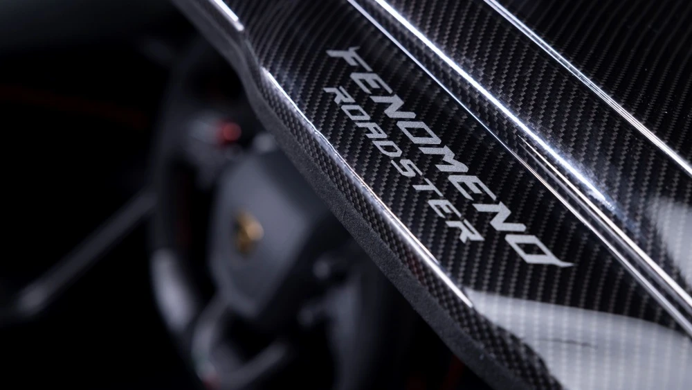 Lamborghini Fenomeno Roadster'ın zarif detaylarını vurgulayan bu yakın çekimde, parlak yüzeyindeki belirgin karbon fiber dokusu ve beyaz harflerle yazılmış FENOMENO ROADSTER ismi dikkat çekiyor; arka plandaki bulanık araç içi öğeler bu yüksek performanslı şasi parçasının modernliğini tamamlıyor.