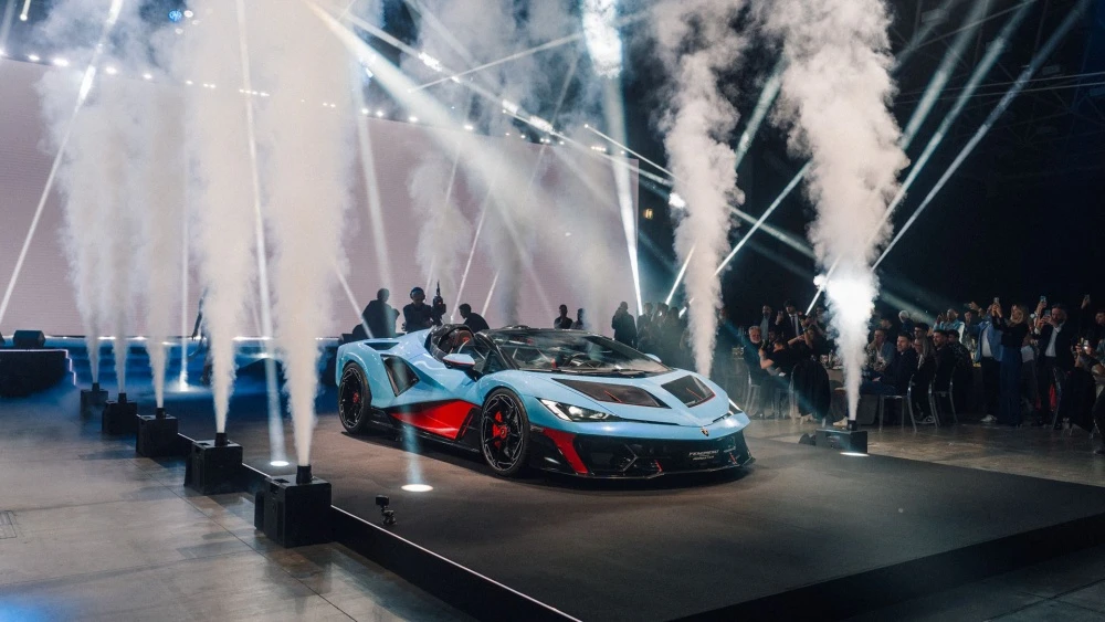 Parlak spot ışıkları ve yükselen duman efektleri altında, açık mavi ve kırmızı detaylara sahip yeni Lamborghini Fenomeno Roadster, lüks bir lansman etkinliğinde şık bir sahnenin merkezinde sergilenirken, heyecanlı izleyiciler bu özel anı cep telefonlarıyla kaydediyor.