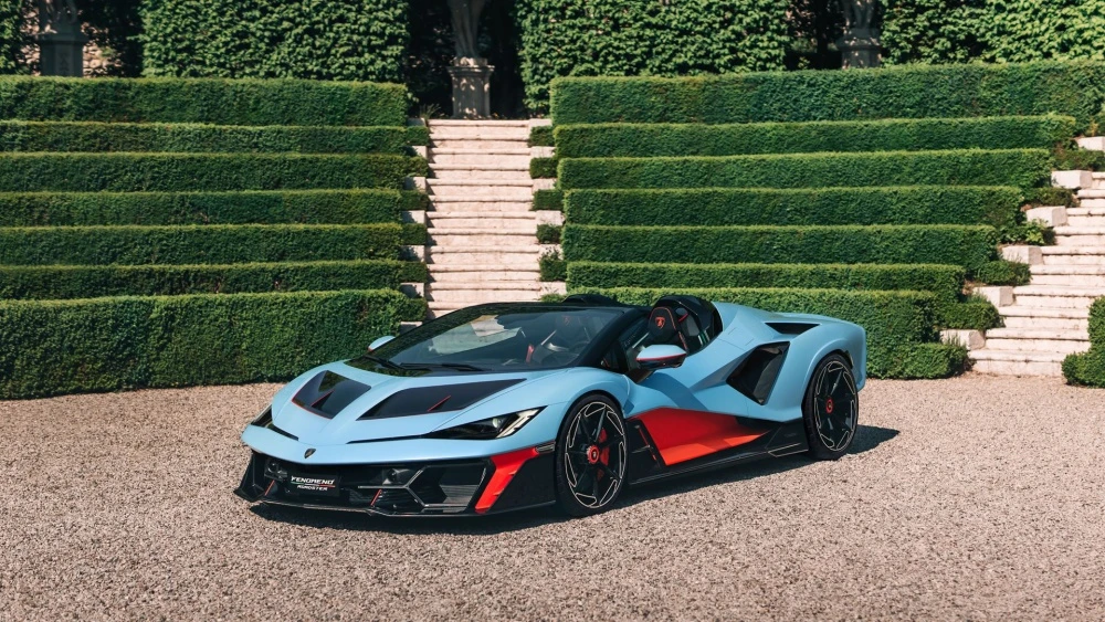 Açık mavi ve turuncu detaylara sahip Lamborghini Fenomeno Roadster, çakıllı bir zeminde, terrasta düzenli çitler ve taş merdivenlerin önünde park etmiş halde, dikkat çekici ön tasarımıyla modern ve agresif bir duruş sergiliyor.