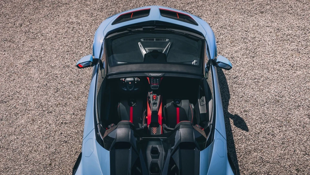 Lamborghini Fenomeno Roadster modelinin üstten çekilmiş bu detaylı görüntüsünde, açık tavanıyla birlikte kırmızı detaylı siyah spor koltukları ve modern tasarımlı kokpiti net bir şekilde ortaya çıkıyor; açık renk çakıl zemin üzerinde park halinde duran parlak açık mavi araç, şık çizgileriyle dikkat çekiyor.