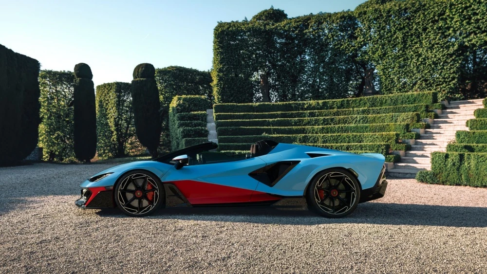 Açık mavi ve kırmızı çift tonlu boyalı, üstü açık bir Lamborghini Fenomeno Roadster, bakımlı yeşil çitlerle bezeli, basamaklı bahçe düzenlemesi olan lüks bir alanda, dinamik yan profil tasarımını vurgulayacak şekilde park edilmiş durumda.