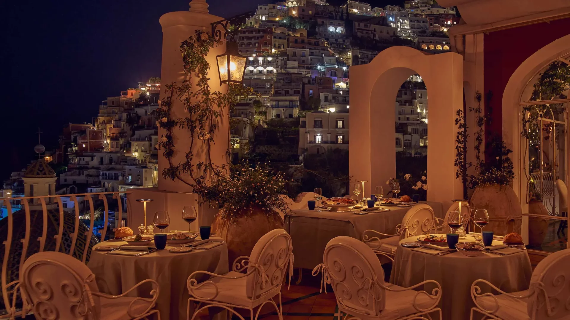 Le Sirenuse restoranının terasından, gece ışıl ışıl parlayan Positano kasabasının manzarasına karşı, zarif beyaz masa örtüleri ve şık sandalyelerle düzenlenmiş akşam yemeği masalarıyla dolu, büyüleyici ve lüks bir yemek ortamı görülüyor.