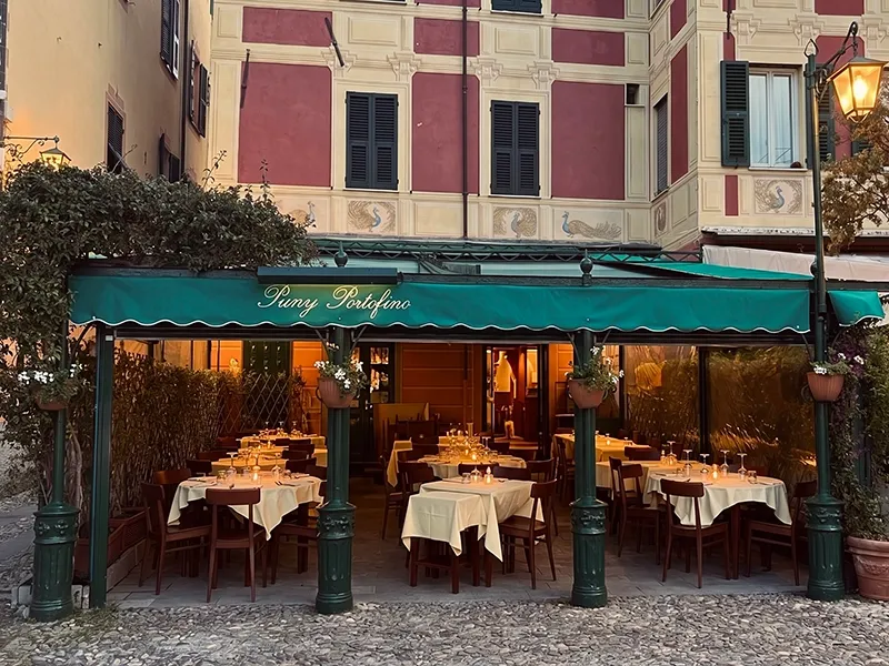 Portofino'da taş döşeli bir meydanda yer alan, yeşil tenteli Puny Portofino restoranının akşam ışıklarıyla aydınlatılmış açık hava masaları, zarif masa düzenlemeleri ve arkadaki binaların duvarlarındaki tavus kuşu motifleriyle sıcak ve davetkar bir Akdeniz ambiyansı sunuyor.
