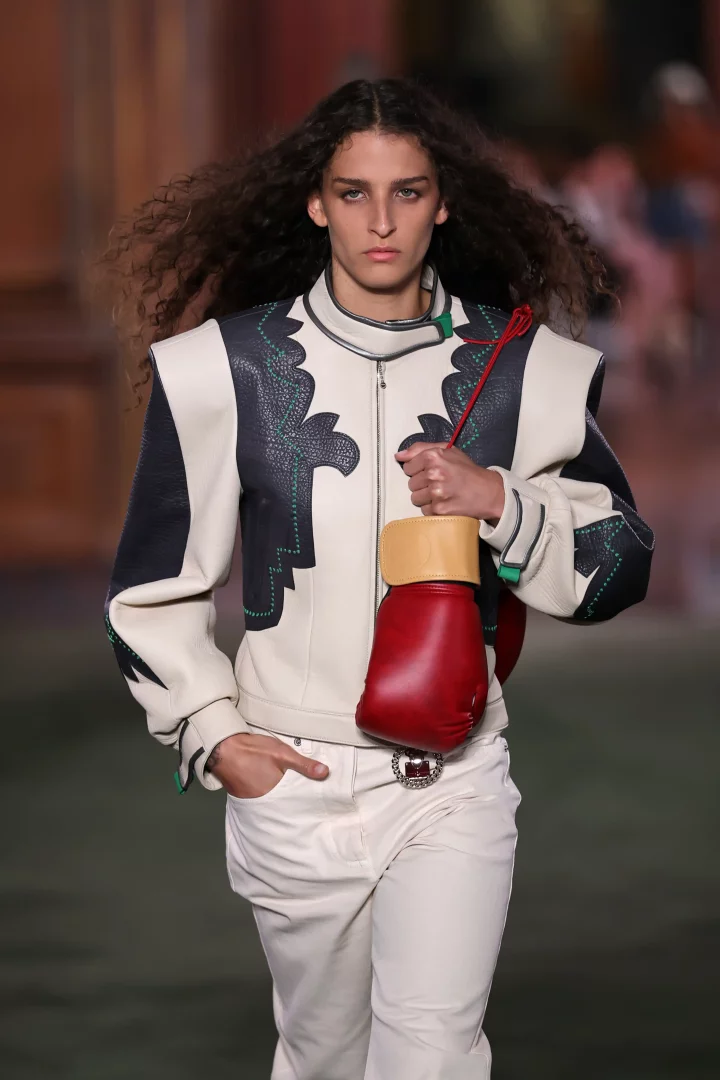 Model, Louis Vuitton Cruise 2027 koleksiyonundan New York'un şehirli ve spor referanslı estetiğini birleştiren, omuzları vurgulu krem rengi bir ceket ve dikkat çekici kırmızı boks eldiveni şeklindeki çantasıyla podyumda ilerliyor.