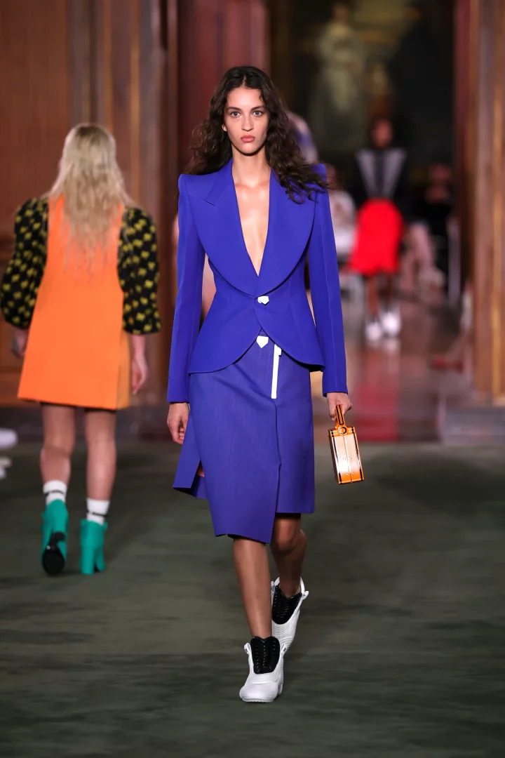 Louis Vuitton Cruise 2027 koleksiyonundan bir manken, The Frick Collection defilesinde, güçlü omuzlara sahip kobalt mavi bir blazer ceket, uyumlu yırtmaç detaylı etek, beyaz spor ayakkabılar ve küçük bir bagaj çanta ile şehirli ve dinamik bir görünüm sergiliyor.