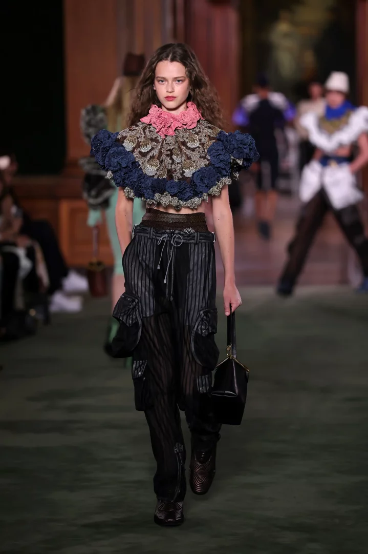 Louis Vuitton Cruise 2027 koleksiyonunun uptown zarafeti ve downtown enerjisini yansıtan bir defile görünümünde, pembe fırfırlı yakalı, işlemeli ve lacivert kabarık omuz detaylı kısa üstü ile bol kesim çizgili kargo pantolonu giymiş model The Frick Collection'ın podyumunda kendine güvenle yürüyor.