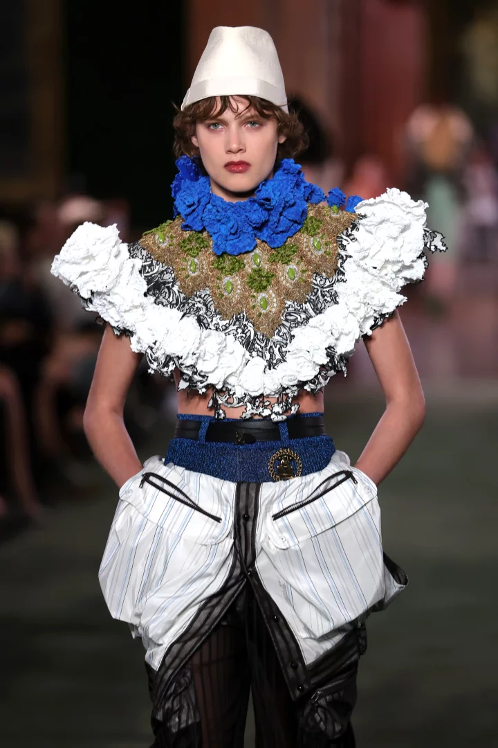 Louis Vuitton Cruise 2027 koleksiyonu defilesinde, beyaz konik şapkası, canlı mavi fırfırlı yakası, işlemeli ve fırfırlı üst kısmı ile dikkat çeken bir model, beyaz çizgili bol cepli ve katmanlı deneysel pantolonuyla New York'un şehirli sanat sahnesinin güçlü bir yansımasını sunuyor.