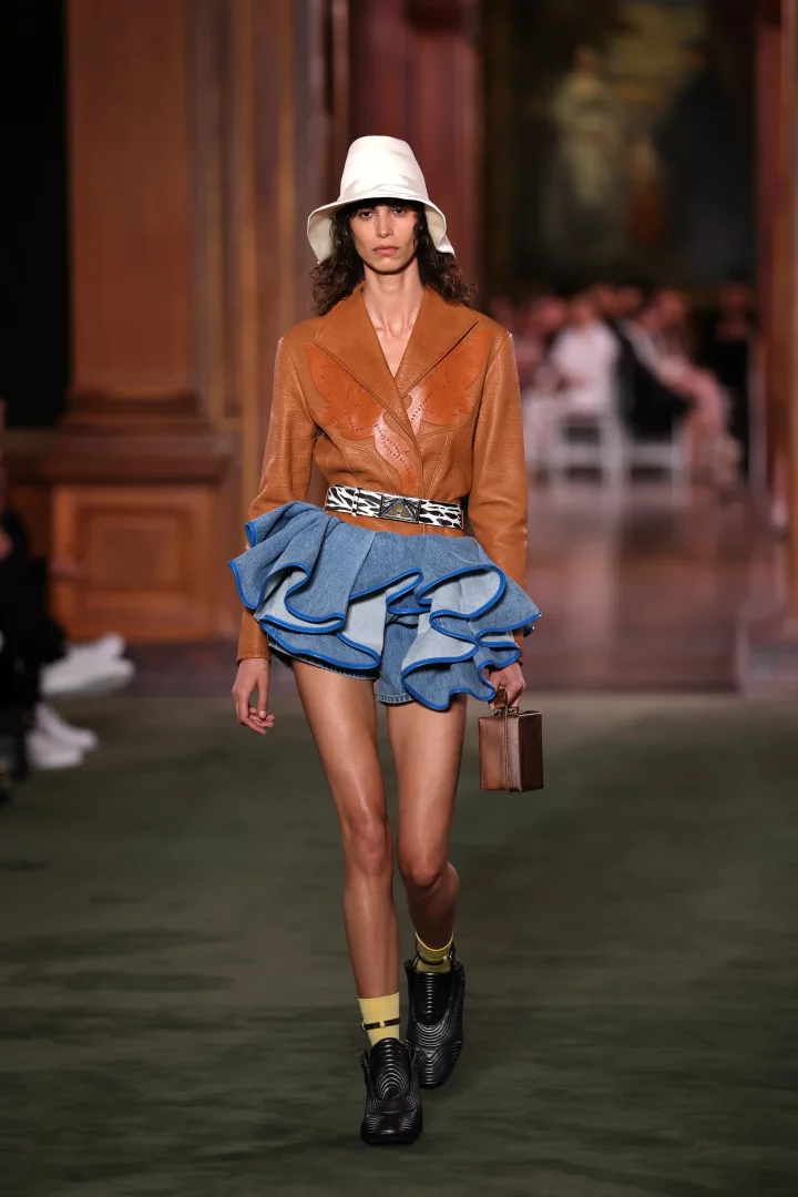 Louis Vuitton Cruise 2027 koleksiyonundan bir görünümle podyumda yürüyen model, kahverengi deri ceketi, katmanlı fırfırlı kot şortu, zebra desenli kemeri ve fütüristik siyah botlarıyla New York'un zarafetini asi ruhuyla birleştiriyor.