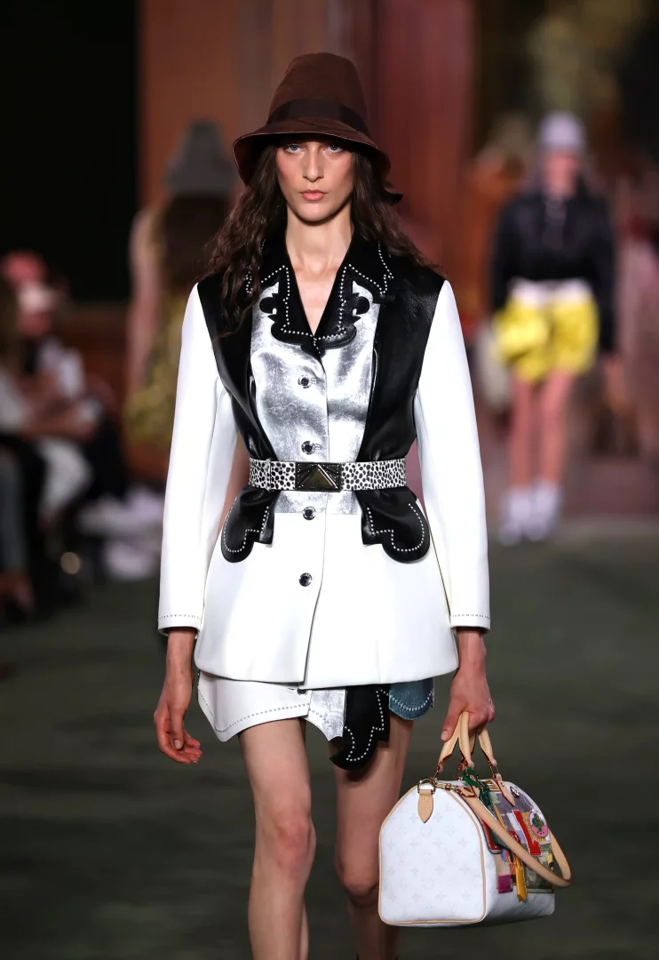 Louis Vuitton Cruise 2027 koleksiyonu için podyumda yürüyen bir model, kahverengi bir şapka, keskin hatlara sahip siyah-beyaz bir ceket, leopar desenli kemer ve sanat baskılı detaylarla süslenmiş ikonik bir Speedy çanta taşıyarak New York'un aristokrat geçmişi ile downtown sanat sahnesi arasındaki kontrastı yansıtan şehirli bir görünüm sergiliyor.