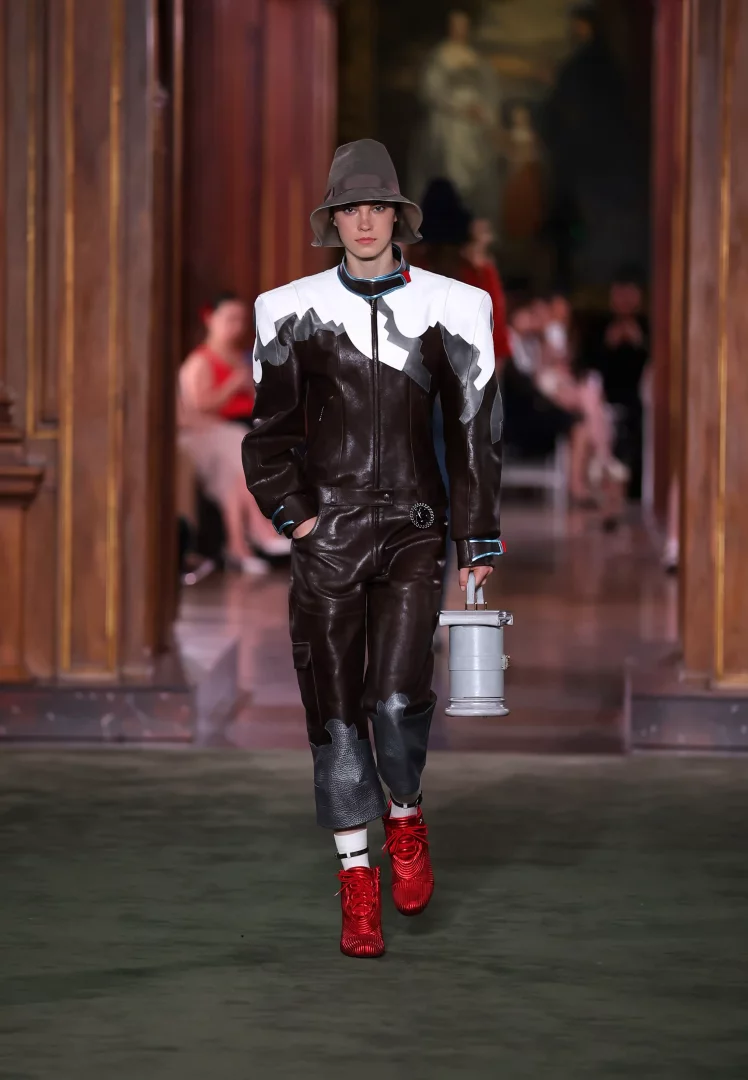 Louis Vuitton Cruise 2027 Koleksiyonu defilesinde, The Frick Collection'ın zengin atmosferinde yürüyen model, güçlü ve heykelsi omuzlara sahip kahverengi deri tulumu, modern gri kova şapkası, parlak kırmızı dokulu spor ayakkabıları ve silindirik gri çantasıyla New York'un sofistike ve şehirli enerji karışımını yansıtan dikkat çekici bir stil sunuyor.