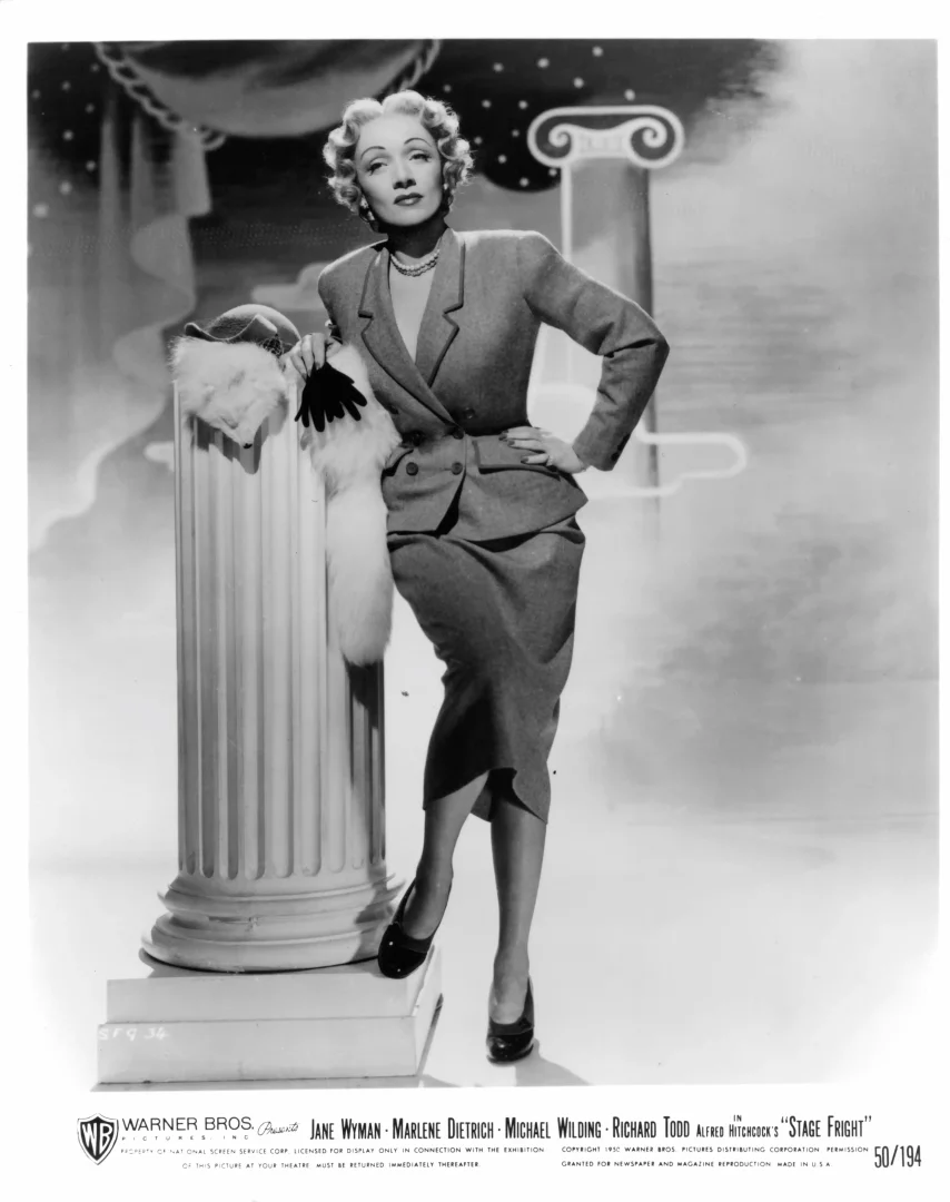 Siyah beyaz fotoğrafta, kıvırcık sarı saçlı Marlene Dietrich, vatkalı, kruvaze bir ceket ve kalem etekten oluşan özel dikim bir takım elbiseyle, beyaz kürklü şal ve eldivenini tutarak klasik bir sütunun yanında zarifçe poz veriyor; bu ikonik görünüm Jonathan Anderson’ın Dior Cruise 2027 koleksiyonu için Hollywood ve Dior arşivlerinden ilham aldığı önemli bir referanstır.