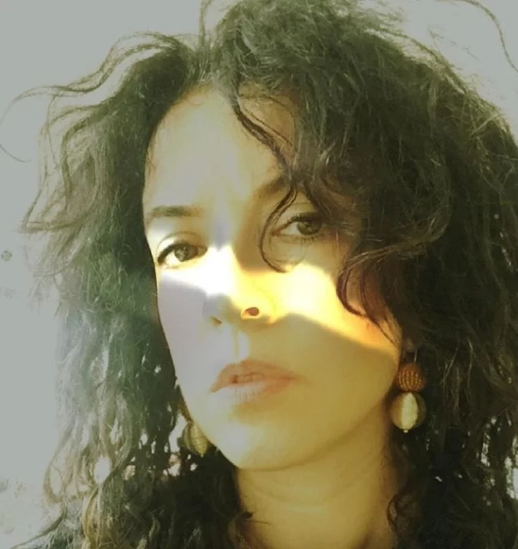 Müge Turan'ın yüzünün sol üst köşesinden gelen güçlü ve sıcak bir ışığın, burnunun üzerinden ve sağ yanağına düşen dramatik gölgelerle birleştiği, koyu kıvırcık saçlarının çerçevelediği, gözlerinin kameraya baktığı sanatsal bir yakın plan portresi.
