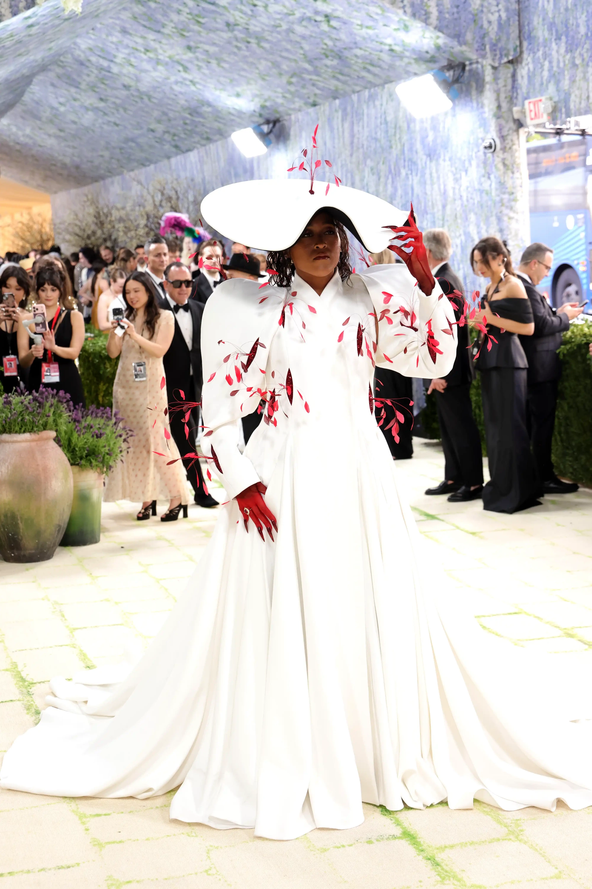 Naomi Osaka, 2026 Met Gala için seçtiği bembeyaz, omuzları vurgulu, üzerinde narçiçeği kırmızısı yaprak motiflerinin uçuştuğu, uzun ve ihtişamlı elbisesiyle dikkat çekiyor; bu iddialı kostümünü geniş kenarlı beyaz bir şapka ve uzun, sivri tırnaklı kırmızı eldivenlerle tamamlayarak Fashion Is Art temasının canlı bir yorumunu sunuyor.